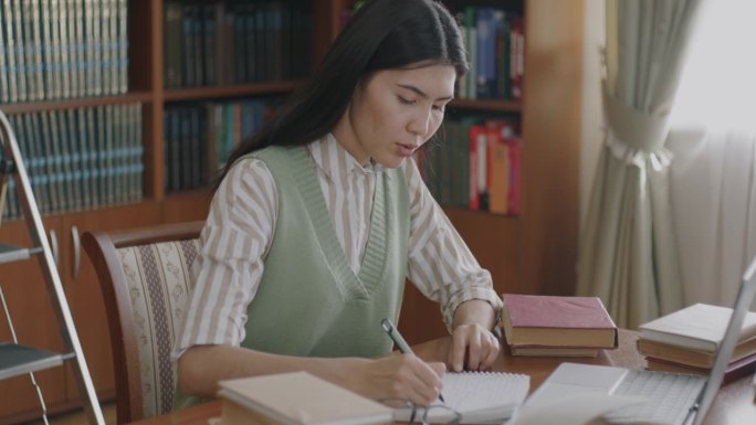 勤奋的亚洲女孩独自在图书馆用笔记本电脑学习，在笔记本上写准备考试