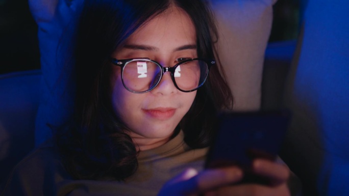 戴眼镜的女人晚上在沙发上玩智能手机