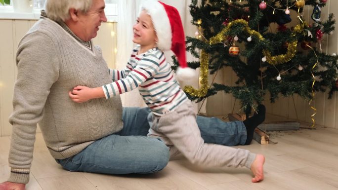 爷爷和小男孩在圣诞节。爷爷和孙子在圣诞树附近的地板上。寒假概念。白发苍苍的老人给蹒跚学步的孩子藏了一