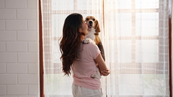 一位年轻的亚洲成年女子站在客厅里抱着她的小猎犬，逗它玩，以示她的爱。