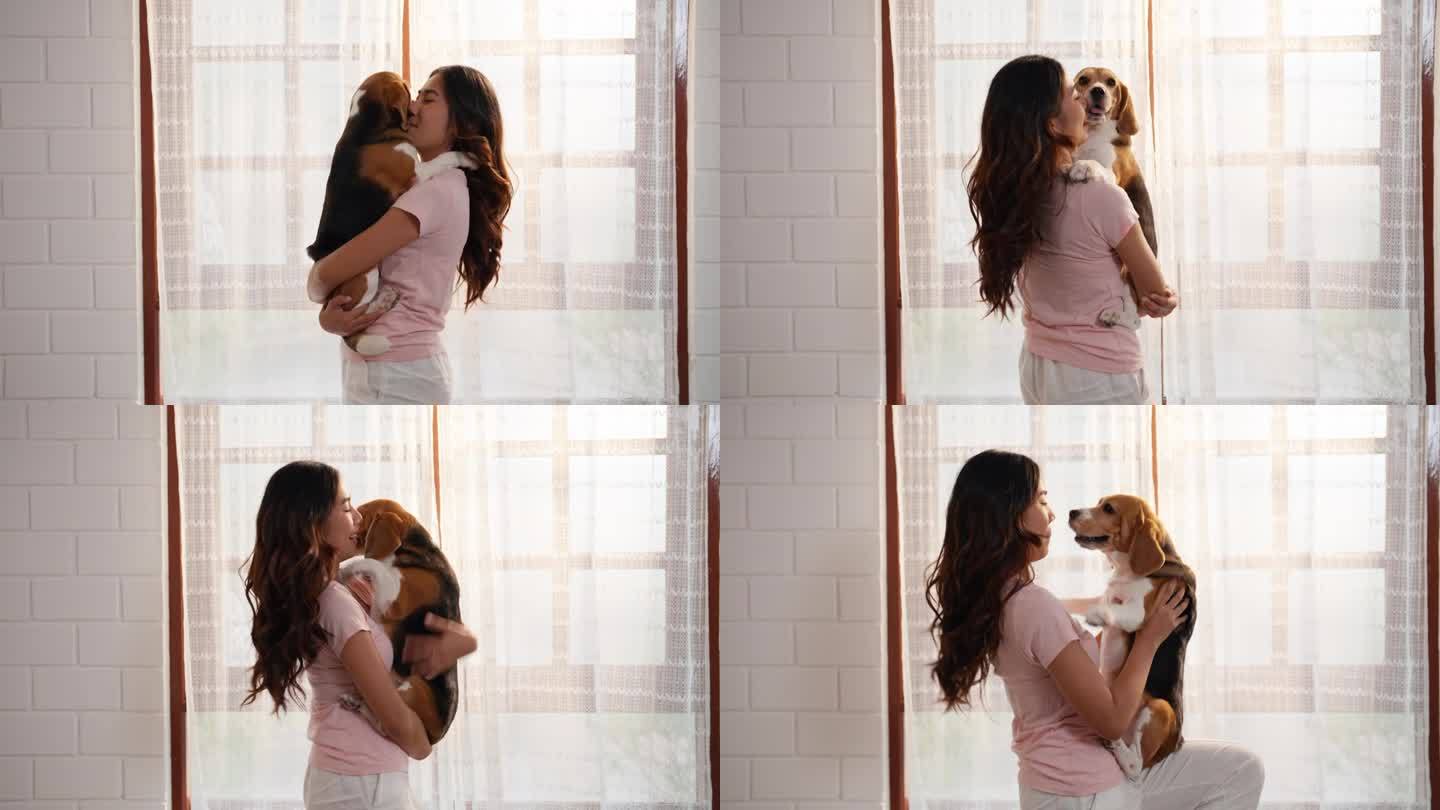 一位年轻的亚洲成年女子站在客厅里抱着她的小猎犬，逗它玩，以示她的爱。