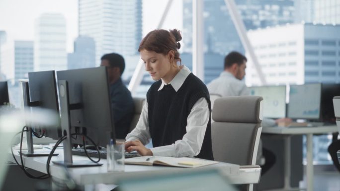 快乐的女客户经理在不同公司办公室的台式电脑上打字，拥有大都市的窗口视图。专业白人女性为咨询公司做笔记