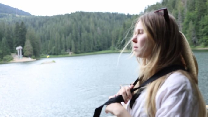 美丽的金发女孩戴着一顶扎着黑丝带的草帽，看着山里的湖。西尼维尔湖附近的一个女人