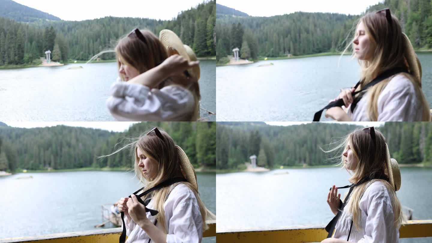 美丽的金发女孩戴着一顶扎着黑丝带的草帽，看着山里的湖。西尼维尔湖附近的一个女人