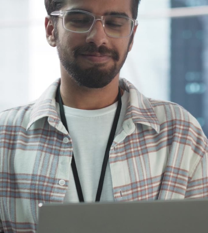 竖屏:英俊的印度男子戴着眼镜，微笑着看着镜头。年轻聪明的男工程师拿着笔记本电脑。现代企业办公室的财务