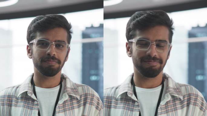 竖屏:英俊的印度男子戴着眼镜，微笑着看着镜头。年轻聪明的男工程师拿着笔记本电脑。现代企业办公室的财务