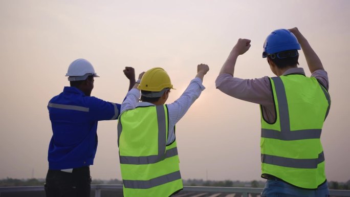 工程师团队在太阳能电池场看日落的后视图。有吸引力的技术工人在屋顶工作，维护和检查光伏电池板系统的太阳