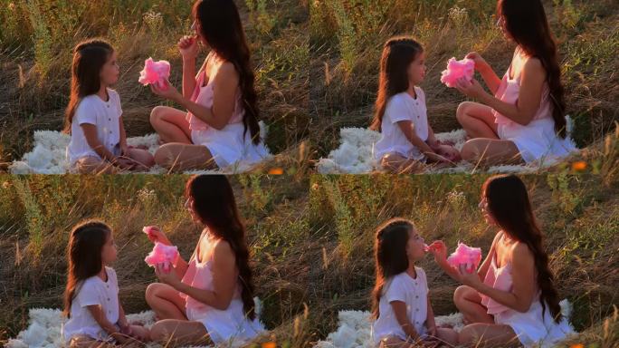 夕阳下，妈妈和女儿在田野里吃粉红色的棉花糖。快乐的童年。度过快乐时光。家庭度假