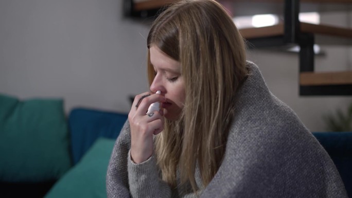 悲伤的疲劳生病的女人擤鼻子喷鼻喷雾剂在慢动作。患病的白种人女士的肖像，流着鼻涕坐在家里接受治疗。