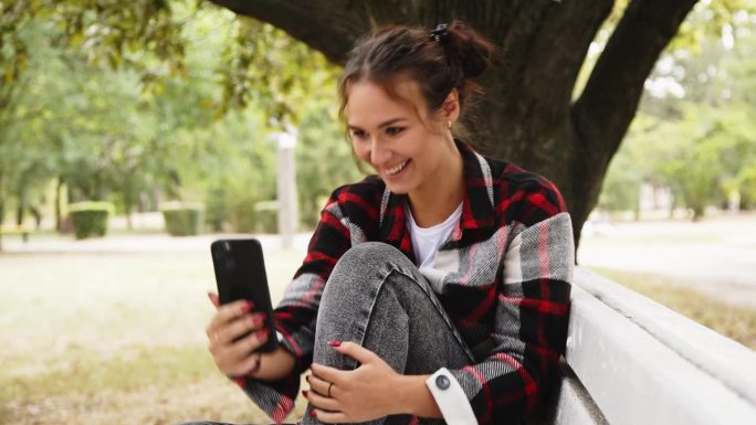 自拍，一个微笑的年轻女子在公园的长椅上用手机拍了一张照片，作为她在社交网络上的快乐回忆。