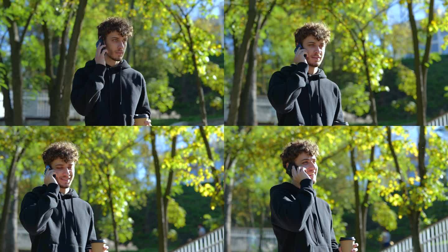 一个面带微笑的年轻人拿着咖啡在公园里散步，一边用智能手机打电话。的生活方式。秋季散步的概念。慢动作