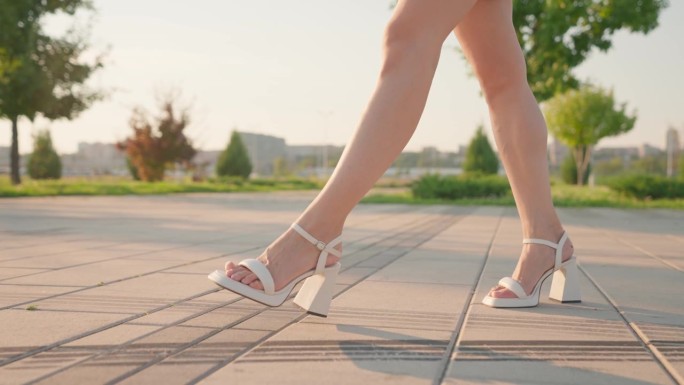 穿着高跟凉鞋的苗条美女在夕阳下漫步城市公园
