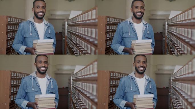 一个聪明的非裔美国年轻人站在图书馆里，拿着一堆书，微笑着享受大学教育