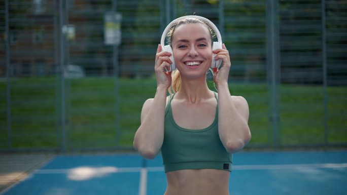 女运动员，跑步者从她的头上摘下白色无线耳机，看着镜头微笑。身材健壮的女孩，穿着漂亮合身的运动服，在镜