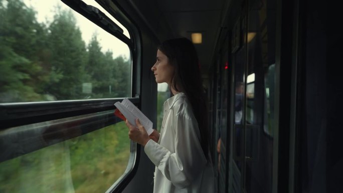 年轻的长发女子或女孩站在快速行驶的客运列车的过道上看书。公共交通工具上没有免费的座位。喜欢坐火车旅行