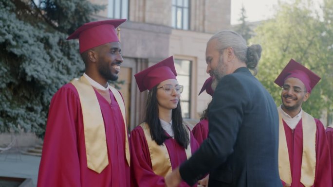 骄傲的教授在大学外祝贺戴着毕业帽、穿着毕业服的不同群体的学生，他们握手拥抱