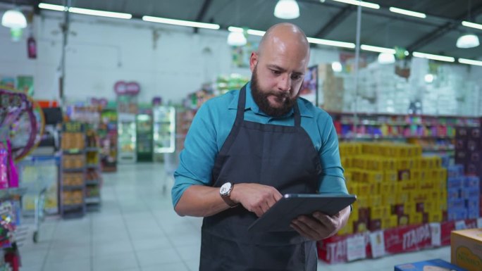 杂货店的巴西员工，戴着围裙，站在连锁超市内，用平板电脑检查库存，专心工作
