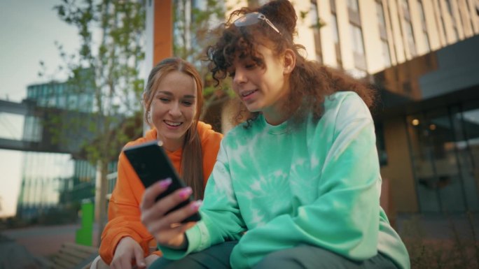 两个年轻时尚的女学生在城市街道上聊天，用智能手机浏览社交媒体帖子。两个时髦的女人在市中心外面讨论时尚