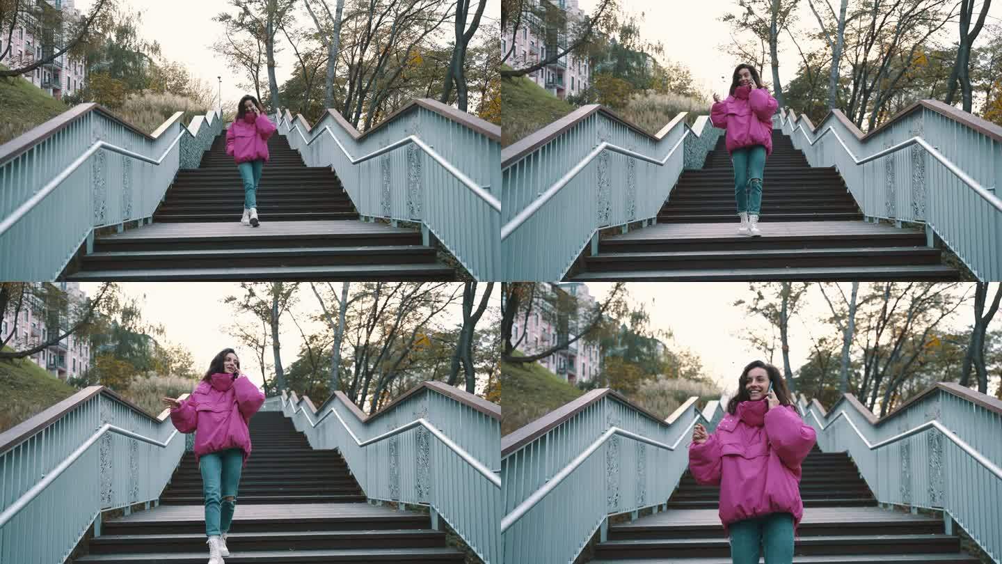 一个穿着时髦夹克、面带微笑的漂亮女人在公园走下楼梯时一边讲着手机。季节性风格，秋季季节概念。慢动作