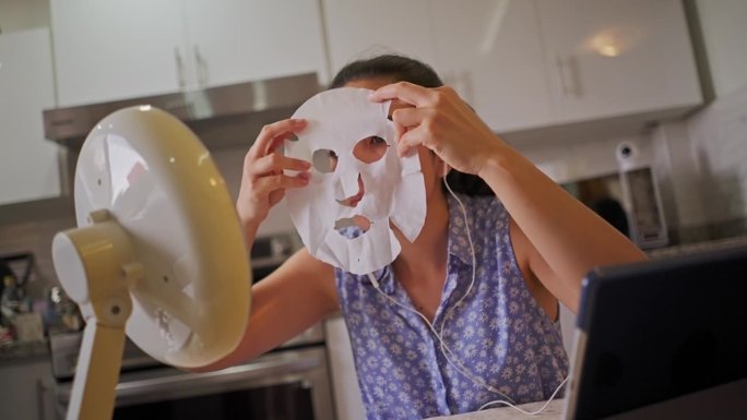 视频通话中的西班牙裔年轻女子一边看着厨房里的镜子，一边给自己敷上保湿、营养、透明质酸和祛痘面膜的慢动