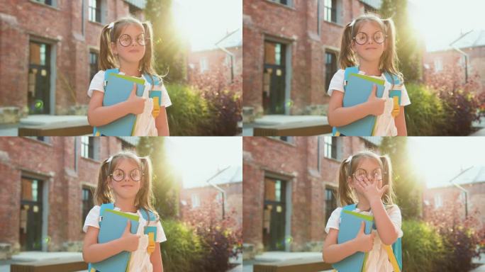 小女孩戴着眼镜，手里拿着背包和书，以学校建筑为背景。快乐的女学生对着镜头微笑。孩子上一年级。