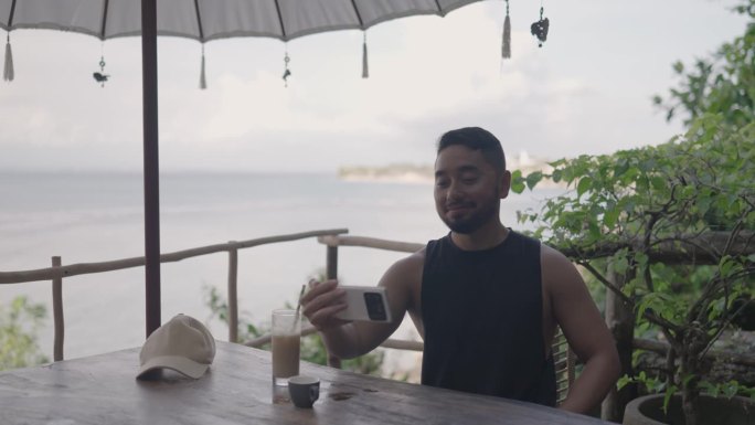 数字游牧民男性男子在印度尼西亚巴厘岛海滩旁的咖啡馆用智能手机进行视频通话