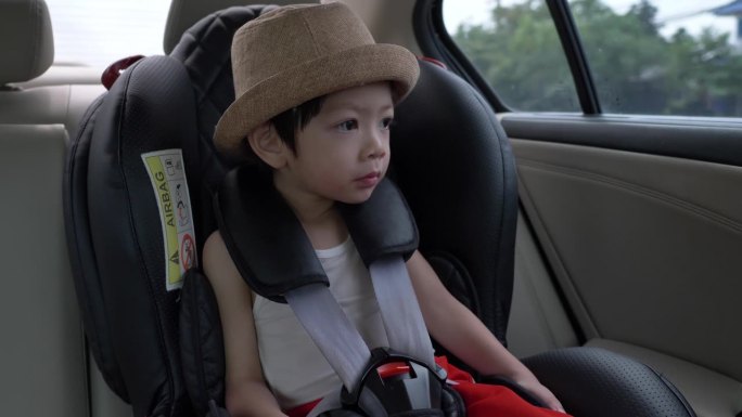 可爱的小男孩戴着帽子坐在安全座椅上，看着窗外的阳光明媚，慢动作的场景