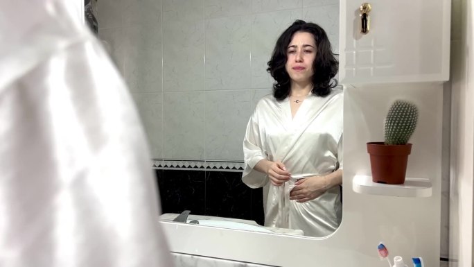 视频中，一位身着长袍的年轻拉丁女性站在家里的镜子前，双手放在肚子上，抱怨自己胃痛。年轻妇女怀孕的人数