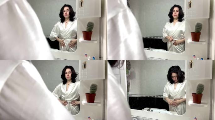 视频中，一位身着长袍的年轻拉丁女性站在家里的镜子前，双手放在肚子上，抱怨自己胃痛。年轻妇女怀孕的人数