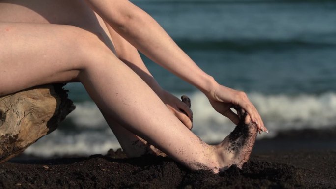 女人光着脚的特写，女人把脚从沙滩上的黑沙子上抖下来，坐在背景波上