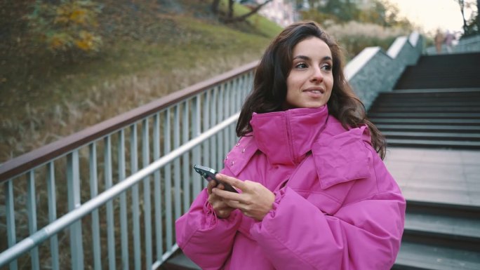微笑的时尚女子站在城市公园里，一边用智能手机打字。秋天的都市街头时尚风格。秋天的季节。实时