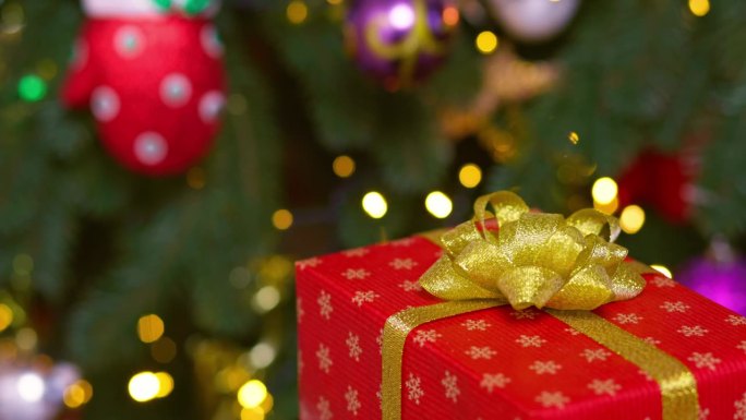 一个系着金色丝带和蝴蝶结的红盒子在闪烁着花环灯的圣诞树的背景下旋转