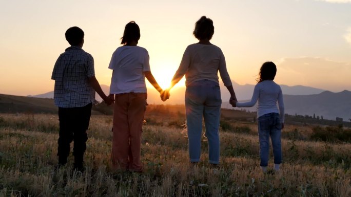妈妈和三个孩子手牵着手站在农村的田野里，看着夕阳的背影。夕阳西下，一家人手拉手走过割过的麦田。友好家