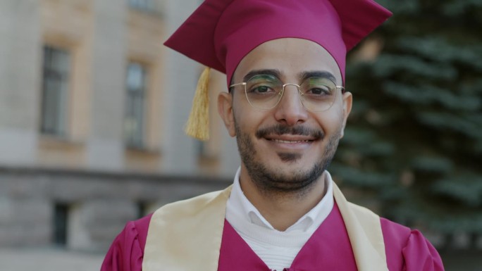 慢动作的肖像，快乐的中东男子学生穿着长袍和学位帽站在大学门前的户外