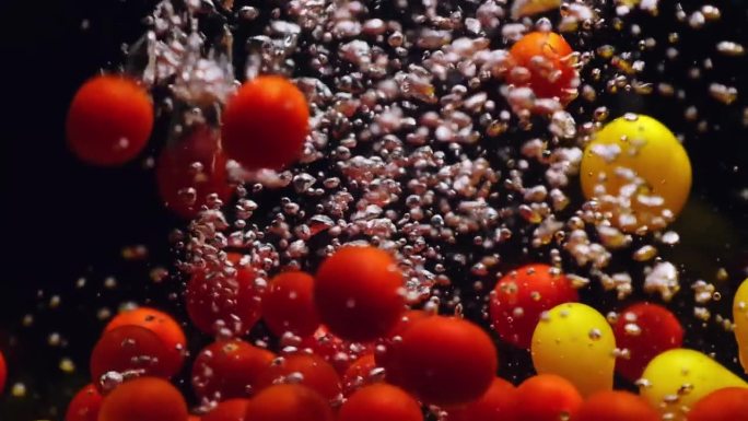 红色和黄色的圣女果落在透明的水中，黑色的背景。各种新鲜成熟的有机蔬菜在水族箱中飞溅，产生气泡。