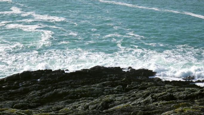 岩石嶙峋的海岸线，背景是广阔的深蓝绿色海洋。暗色的岩石参差不齐，在无情的海浪冲击下屹立不倒，形成白色