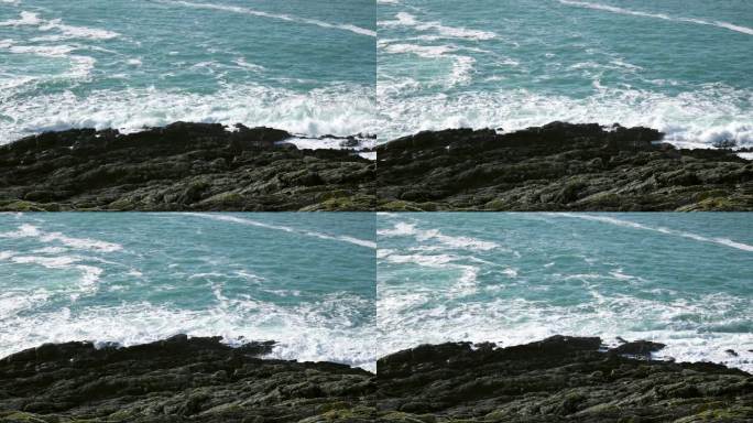 岩石嶙峋的海岸线，背景是广阔的深蓝绿色海洋。暗色的岩石参差不齐，在无情的海浪冲击下屹立不倒，形成白色