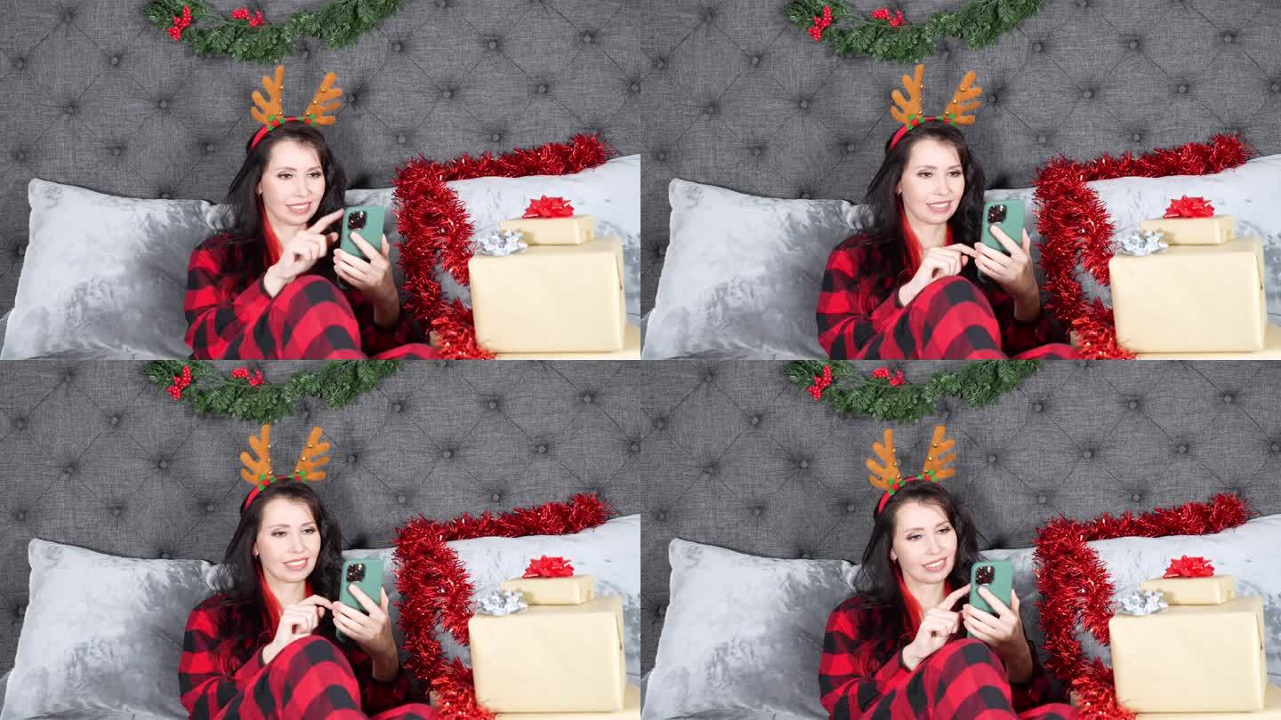 年轻漂亮的女人穿着红色格子睡衣，在摆满礼物的圣诞装饰房间里滑动着智能手机，微笑着。
