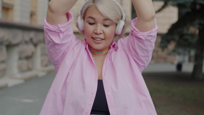 活跃的亚洲女孩戴着耳机听音乐，在城市街道外跳舞的慢动作