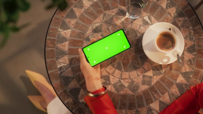 俯视图的匿名女性使用智能手机模拟绿屏Chromakey显示，而坐在咖啡桌。优雅女子横着拿着手机看视频