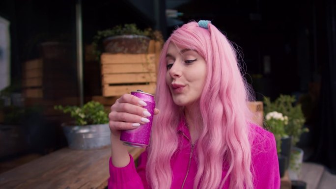 一个女孩穿着粉红色的衣服和粉红色的头发在街上喝苏打水从一个粉红色的罐子。粉红色色彩和谐，个性鲜明