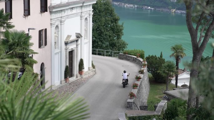 一个人骑着摩托车经过一座古老的教堂，身后是绿色的湖