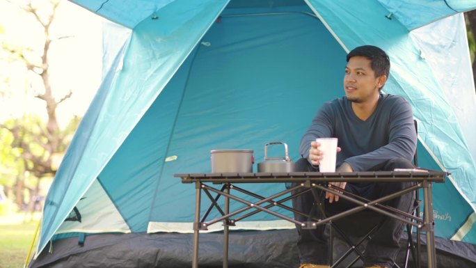 一名亚洲男子坐在露营帐篷前休息，一边喝着热咖啡，一边在森林露营地放松。