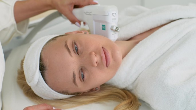 漂亮的女性客户穿着浴袍躺在美容床上的脸部特写，用超声波装置进行颈部提拉手术。