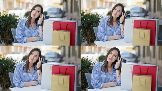 年轻美丽的西班牙女子坐在咖啡店露台上拿着购物袋的桌子上，用智能手机听语音信息
