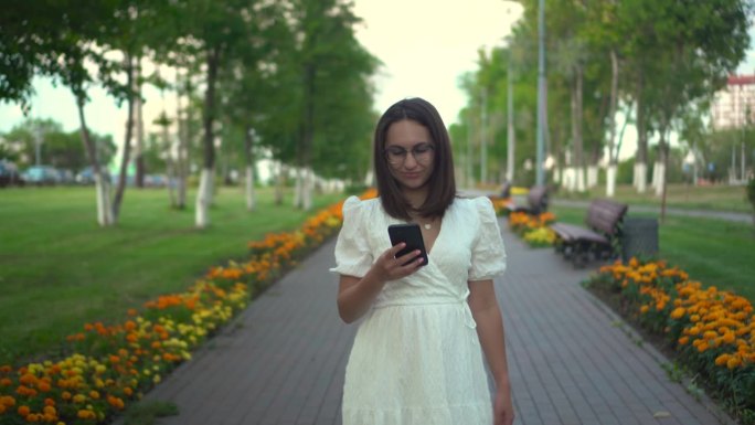 一位年轻女子手里拿着电话走在花巷里。一个穿白裙子的女孩看着她的手机，走在花丛中。4 k