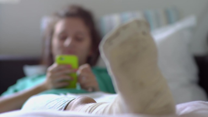 脚部骨折的年轻女子在脚部受伤事故后躺在家里的沙发上，用智能手机上网