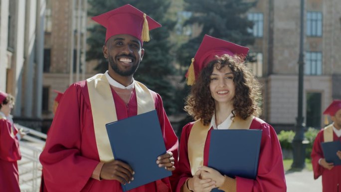 非裔美国男性和白人女性毕业生的肖像站在校园里，拿着学士学位文凭，戴着迫击炮板