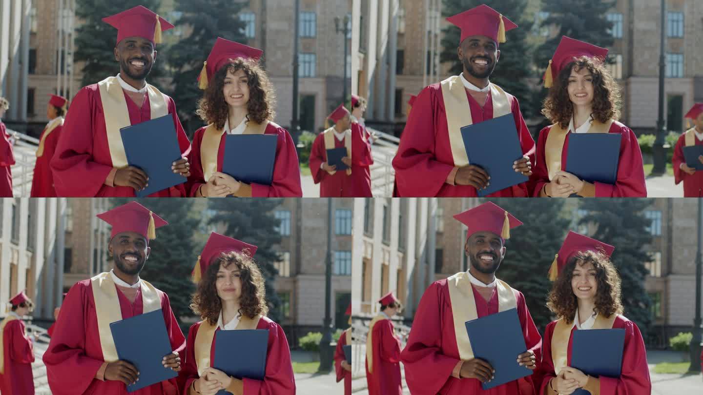 非裔美国男性和白人女性毕业生的肖像站在校园里，拿着学士学位文凭，戴着迫击炮板