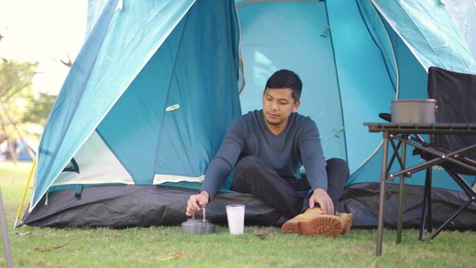 一名亚洲男子坐在露营帐篷前休息，一边喝着热咖啡，一边在森林露营地放松。